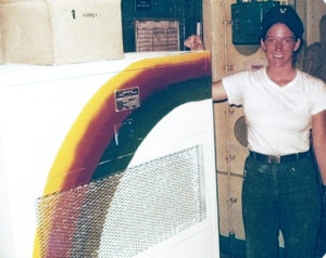 Sharon in uniform T-Shirt, dungarees and cap with Naval insignia standing beside an AC unit painted white with a rainbow. Aboard the USS Jason circa 1981